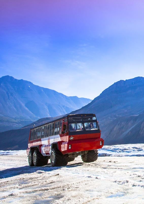 A red and white Ice Explorer drives down a glacier before a blue and purple sky.