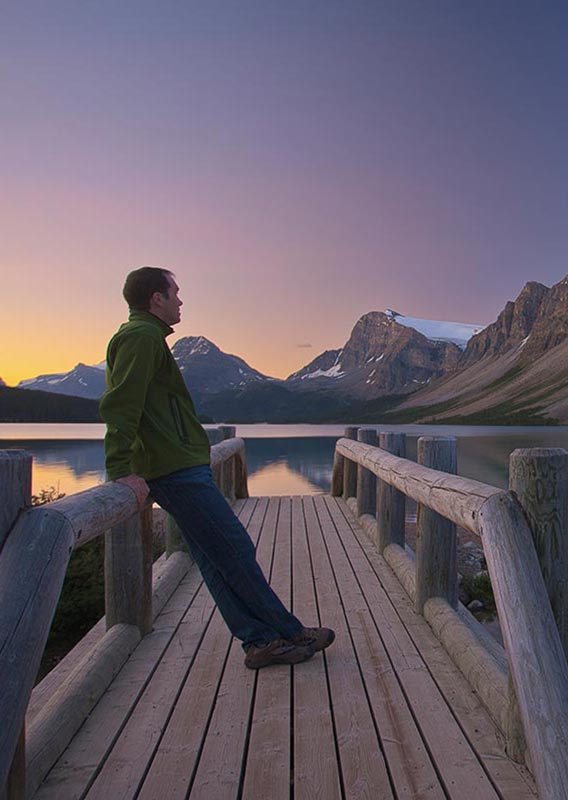 A man stands on a small wooden footbridge looking out to a mountain across a calm lake.