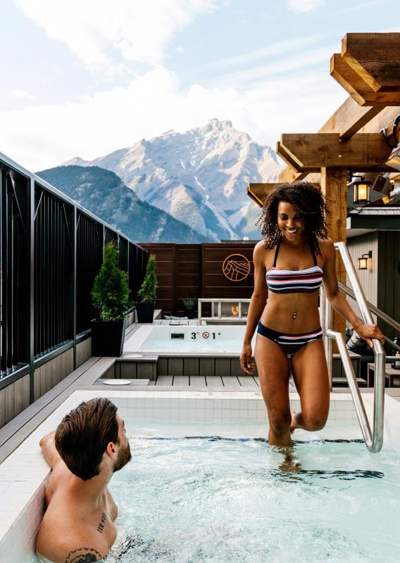 Two people relax in a rooftop hot tub with views of mountains in the distance.