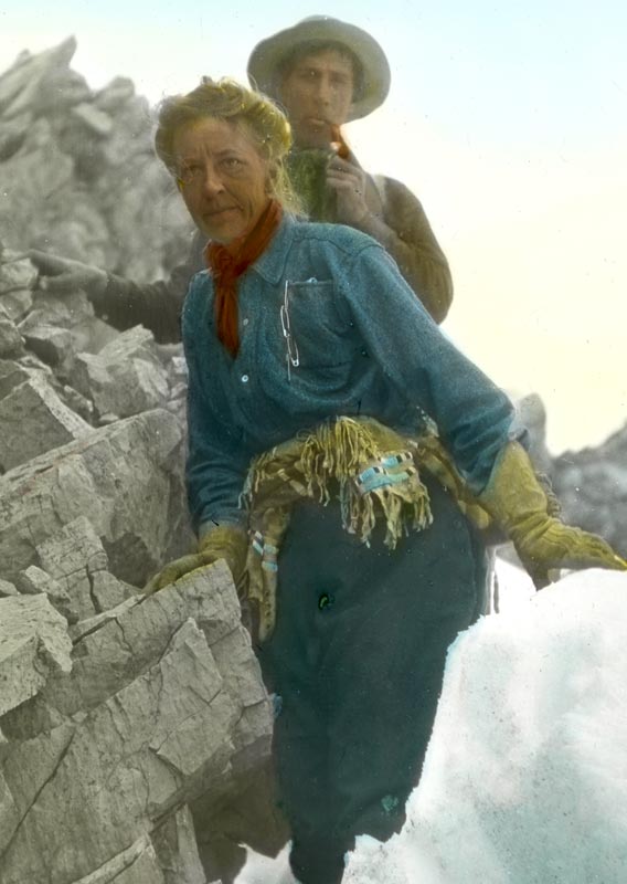A woman hikes between rocks and snow, followed closely by a man smoking a pipe