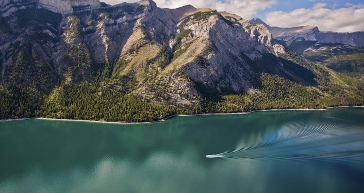 An aerial view of a boat cruising along a large blue-green lake below tree-covered mountains.