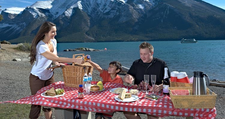 A family sits at a picnic table next to a large lake with a spread of food and drinks.