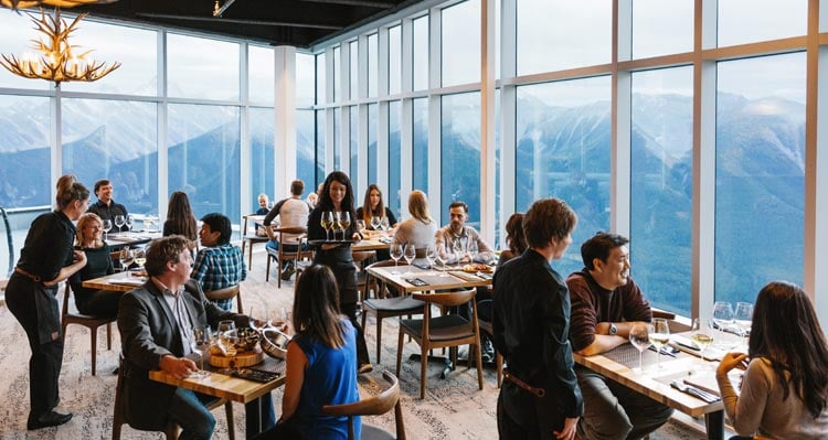 Busy restaurant tables at Sky Bistro, with floor-to-ceiling windows revealing wide mountain views.