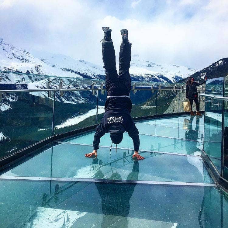 A person does a handstand on a glass walkway, overlooking a wide, snowy valley.