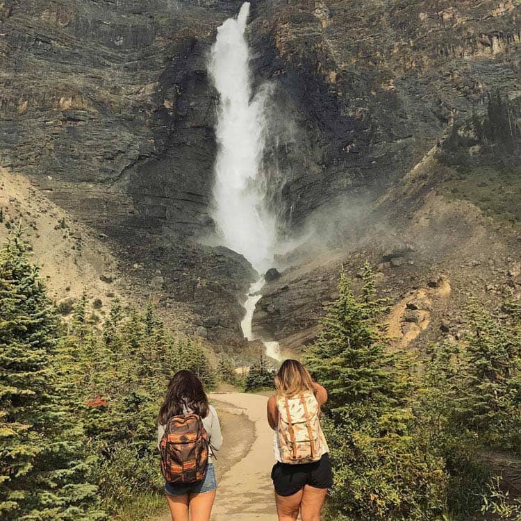 Two hikers stands at a misty waterfall by rocky cliffs.