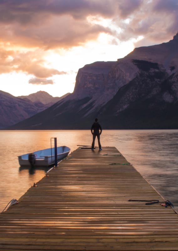 A person stands at the end of a dock overlooking a calm lake surrounded by mountains.