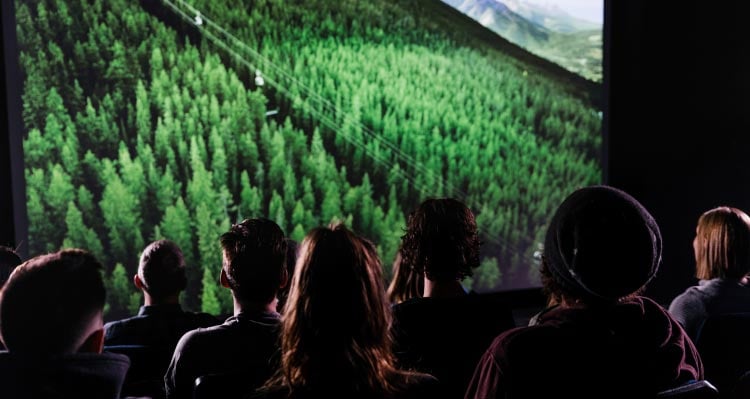 A group of people sit in a theatre watch a film showing the Banff Gondola on a green mountainside.