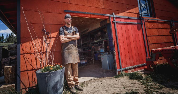 A blacksmith stands outside a red barn workshop.