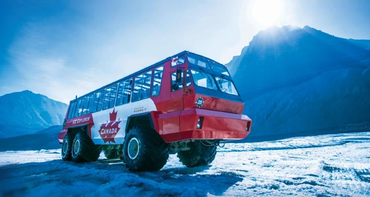 A red and white Ice Explorer vehicle drives on a glacier with sun glowing behind a mountain.