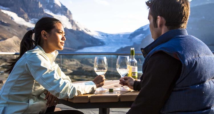 Two people enjoy a bottle of wine at a table overlooking a glacier.