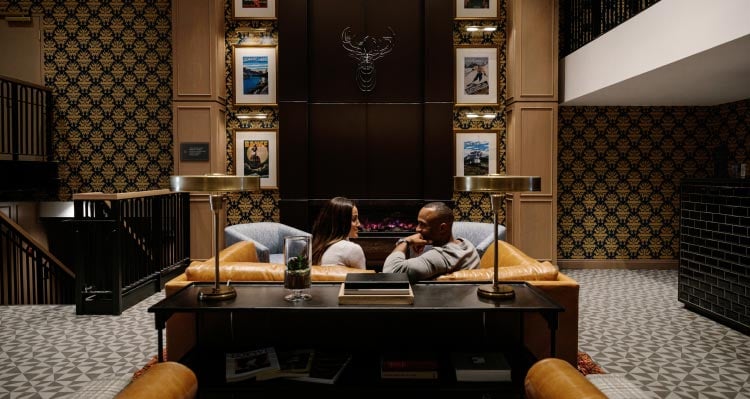 Two people sit in a leather couch in a tall lobby with artwork above a fireplace.