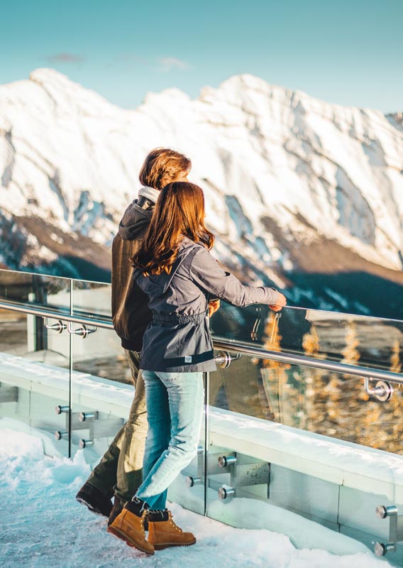 Couple standing at a mountaintop railing looking towards snow-covered peaks.