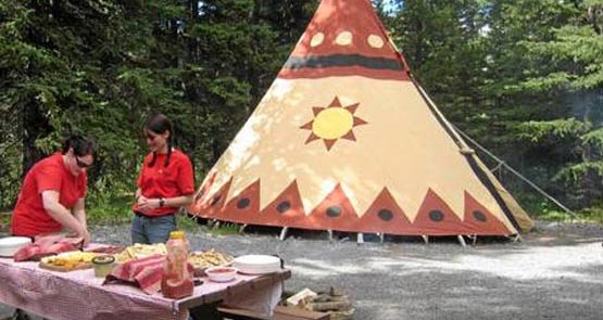 Two museum workers stand at a table outside of a tipi.