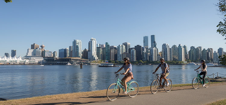 People ride bikes on a shoreline path with the city skyline in the background