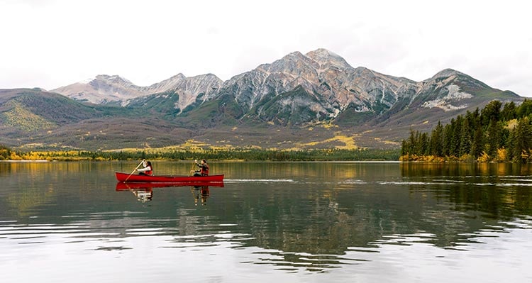 Two people in a canoe on a calm lake below a mountainside.
