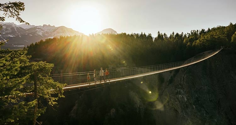 A family walks across a long suspension bridge with the sun shining from behind mountains.
