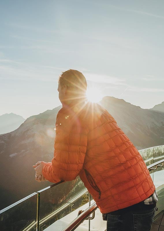 A person in an orange jacket stands at a balcony railing looking towards a mountain.