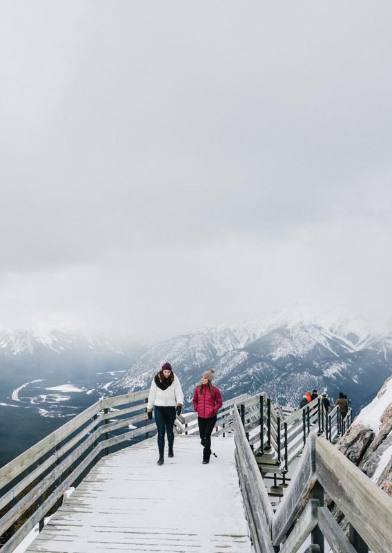 Two people walk along a mountaintop boardwalk above a snowy valley.