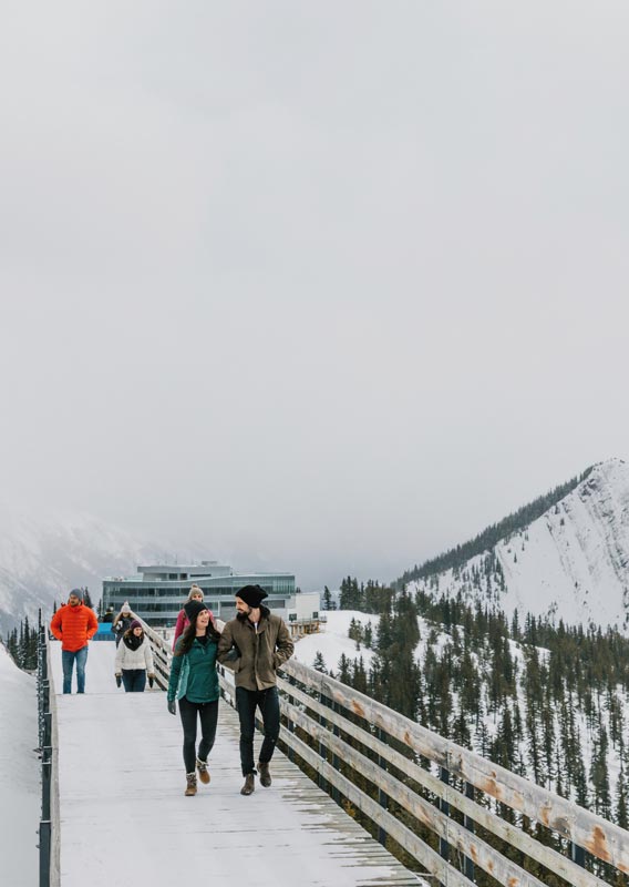 Groups of people walk on the Sulphur Mountain boardwalk among snowy peaks.