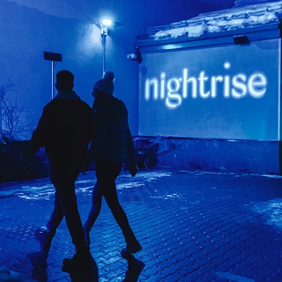 Two people walk past a lit sign for Nightrise.