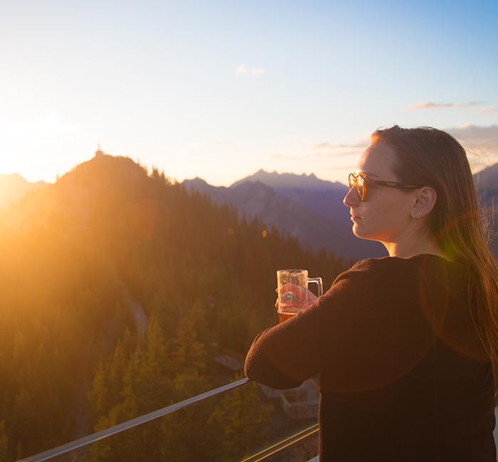 A woman stands at a balcony railing overlooking a tree-covered mountain and the sunset.