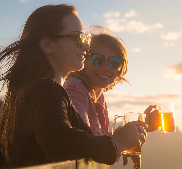 Two women hold drinks and look over a barrier at the sunset