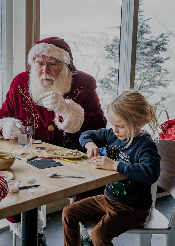 Santa sits at a table with children decorating cookies