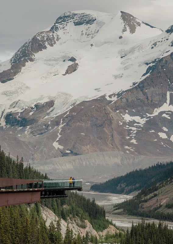 A metal and glass platform extends out over a valley below a tall glacier-covered mountain.
