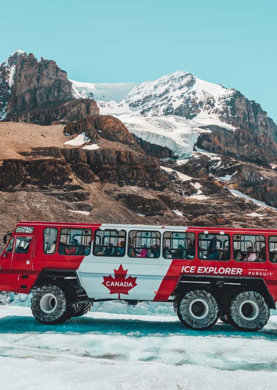 A red and white Ice Explorer bus drives along a glacier below a glacier covered mountain.