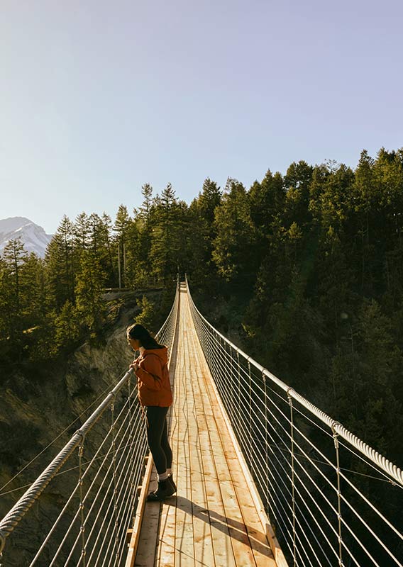 Two people walk across a wooden suspension bridge over a forested canyon.