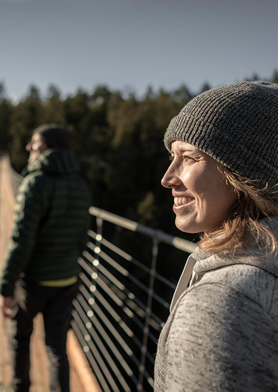 A person looks out from a wooden suspension bridge across a forested canyon.