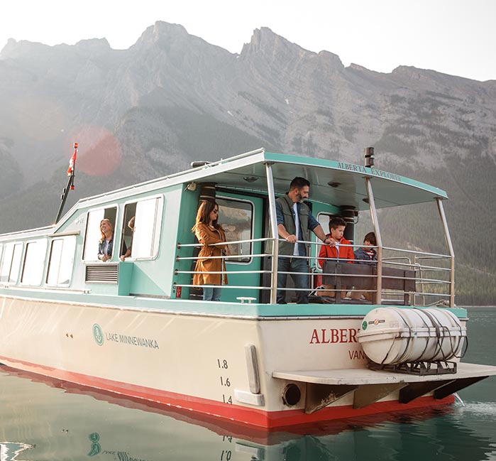 A family stands at the back deck of a turquoise and white boat on a lake below mountains.