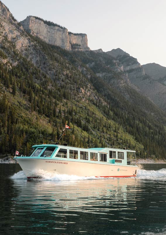 A blue and white boat cruises on a lake below a forested mountainside