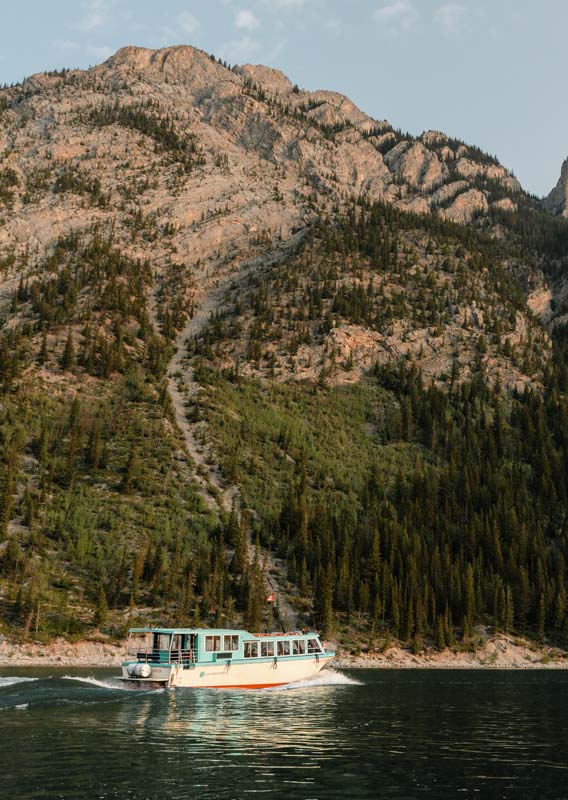 A blue and white boat cruises on a lake below a forested mountainside