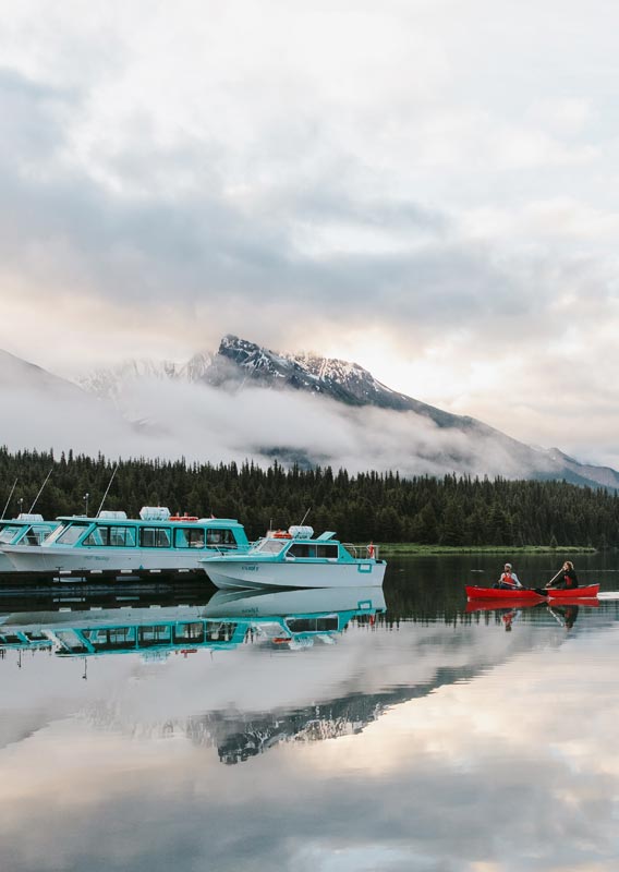 A boat cruises across a lake beneath tree and snow-covered mountains