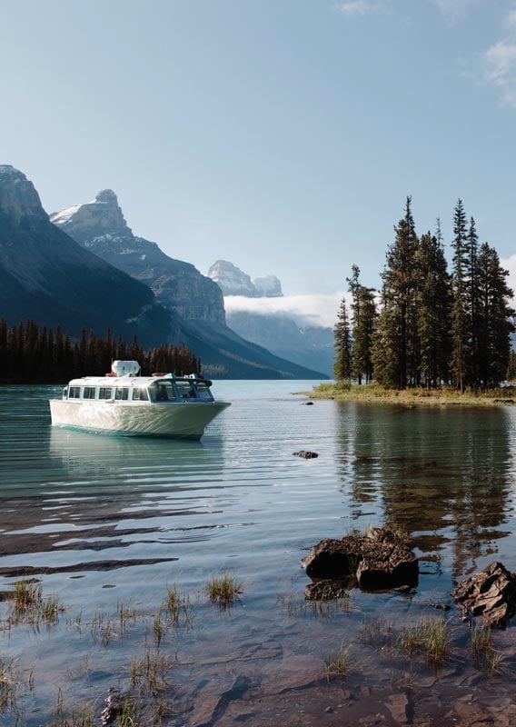A boat set in a calm lake surrounded by mountains with a grove of trees on a small peninsula nearby.