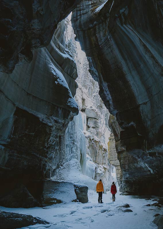 Two people stand at the frozen bottom of Maligne Canyon in front of frozen waterfall