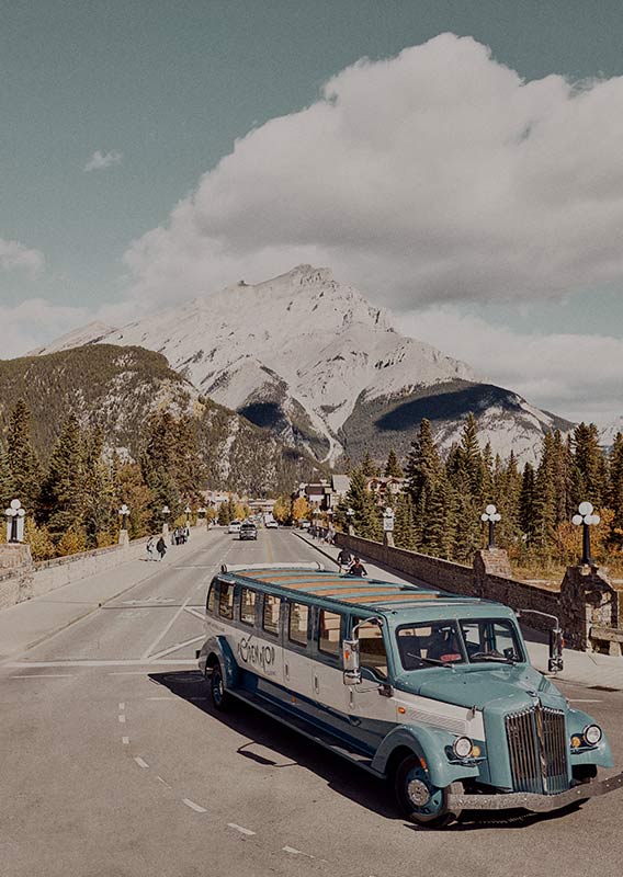 A historic style bus drives from a bridge away from a small town, with mountains in the background.