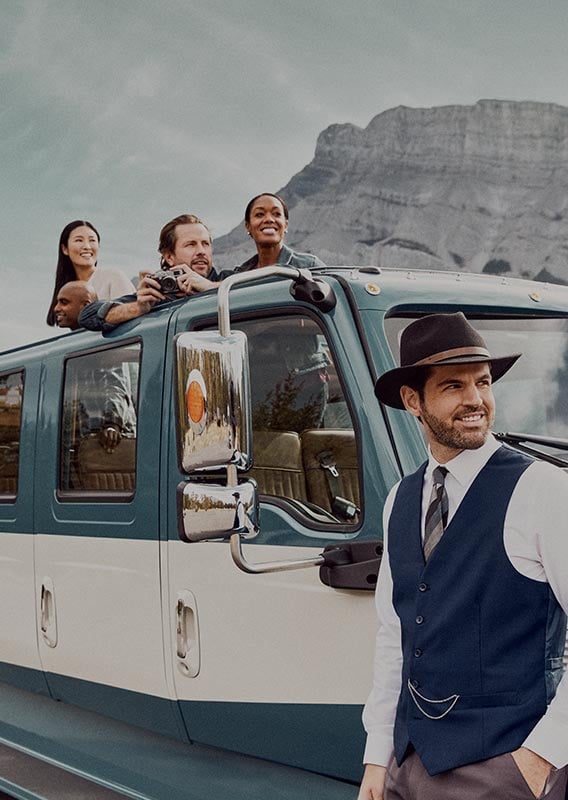 A driver stands out front of a historic-style bus with a group of people looking out from the roof.