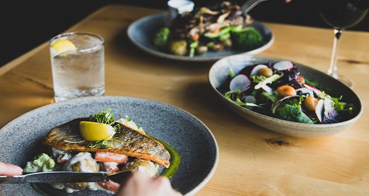 Plates of various food in the middle of a wooden table, glasses of water and wine beside.