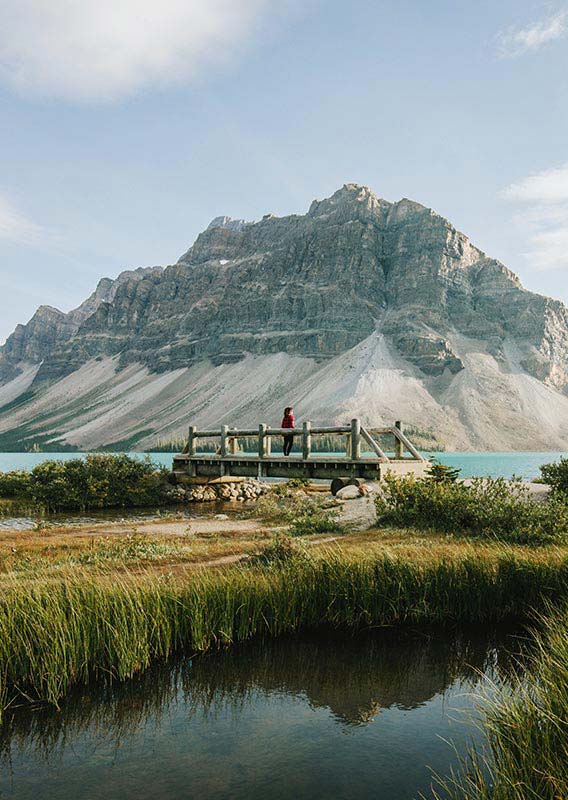 A person stands on a wooden bridge alongside a blue lake and soaring mountain.