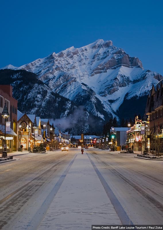 A dusk-hour view down a wintry street towards a tall snow-covered mountain.