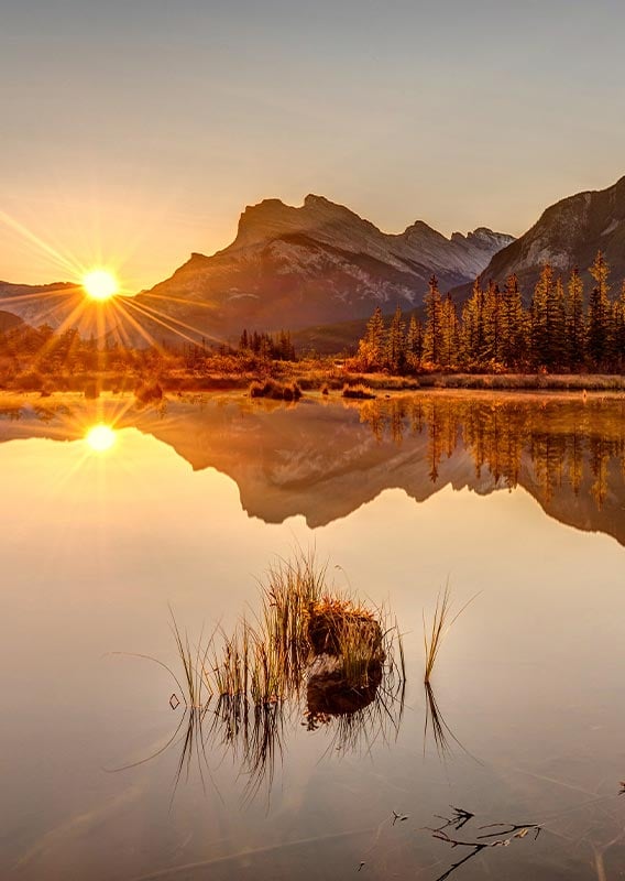 The sun rises from behind a horizon of mountains, shining off of a calm lake.