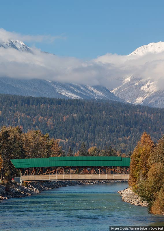 A green covered bridge stretches over a blue river beneath forested mountainsides.