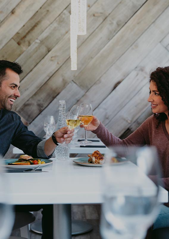 Two people sit at dinner table raising wine glasses for a cheers.