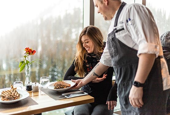 A chef brings a dinner plate to a diner at a windowside seat.