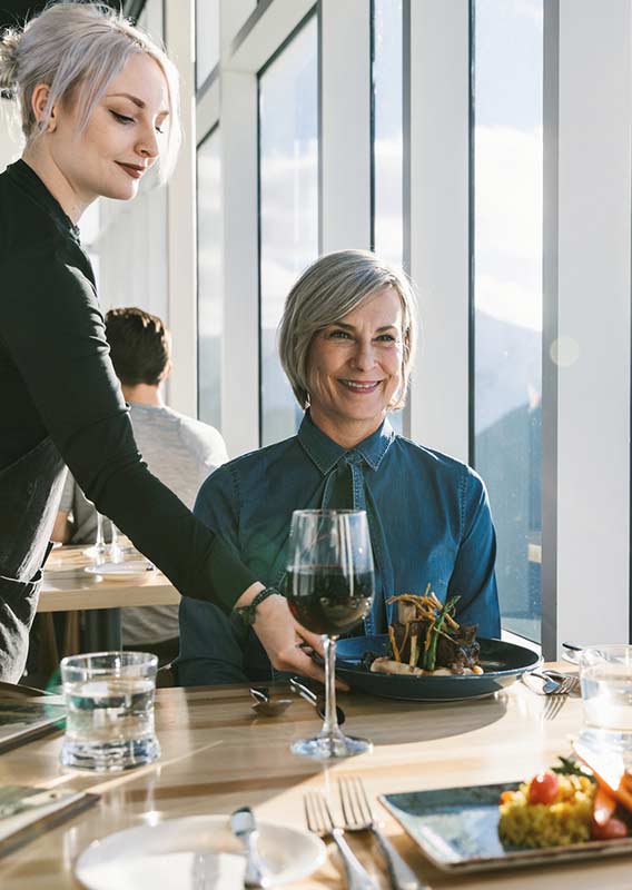 A server brings a dinner plate to a table at a windowside.