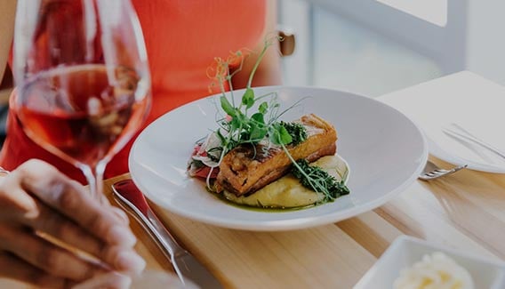 A plate of pork belly and polenta in front of a person holding a wine glass.