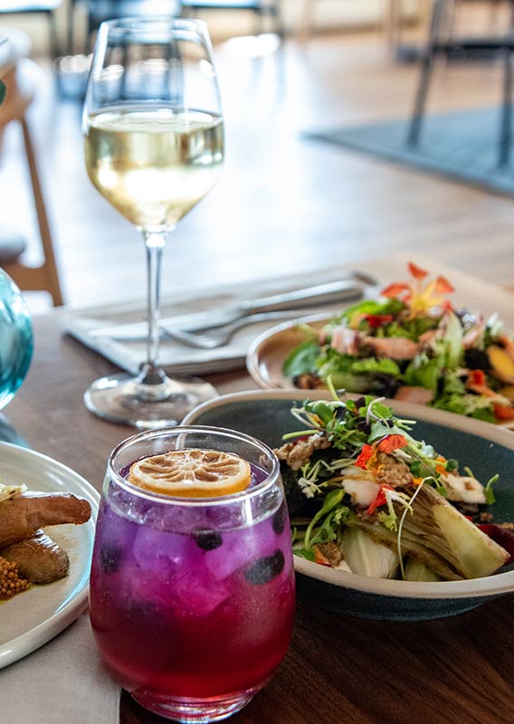 A table of meals featuring a glass of white wine a colorful cocktail.