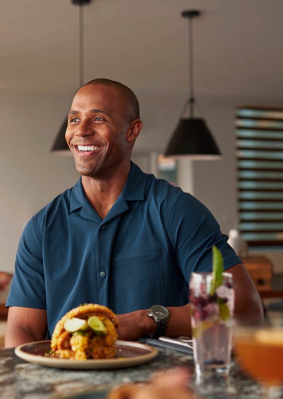 A man is smiling at a dinner table, with food and drink in front of him.